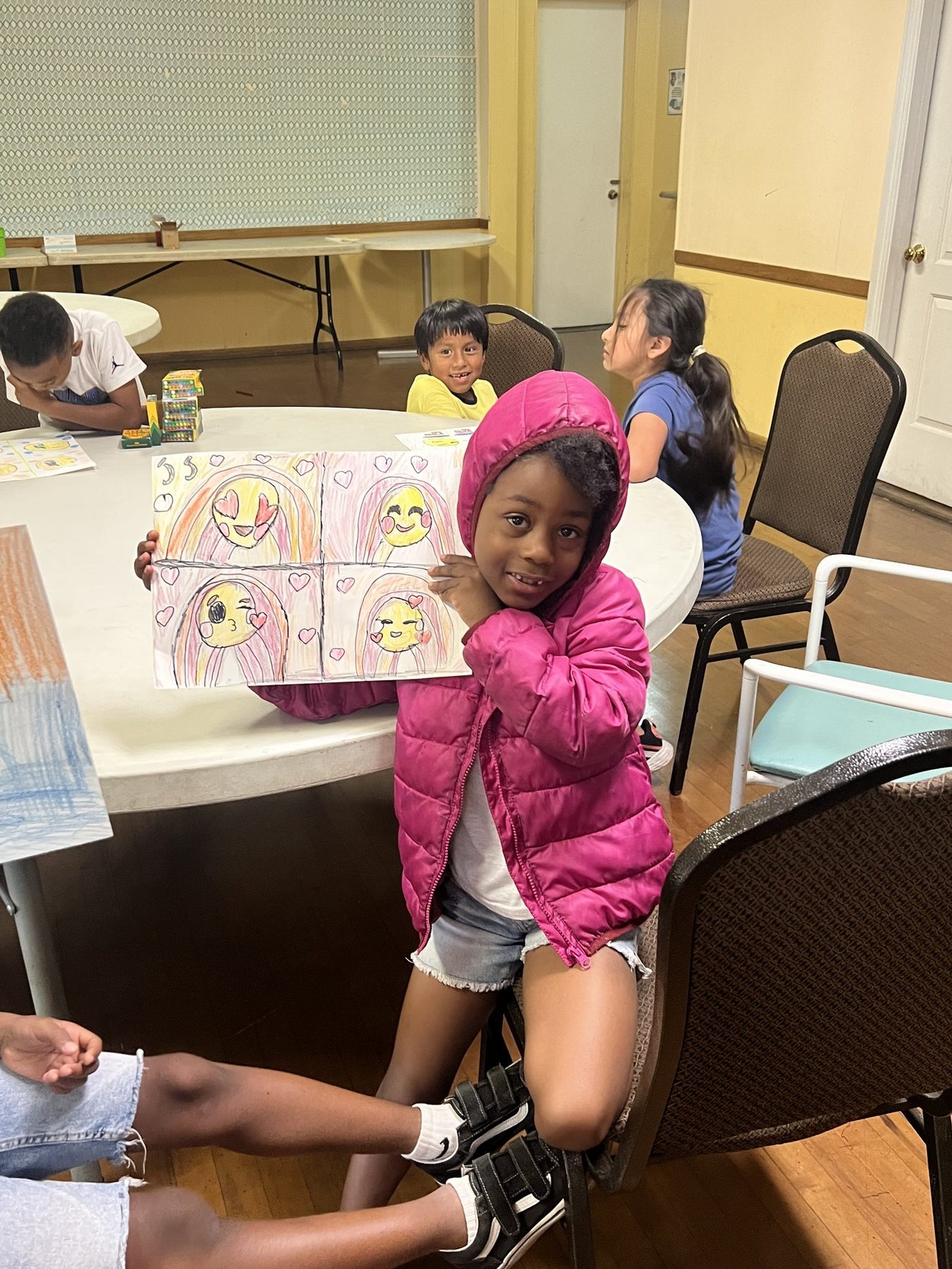 A student smiling and holding up her emoji drawings at the art table